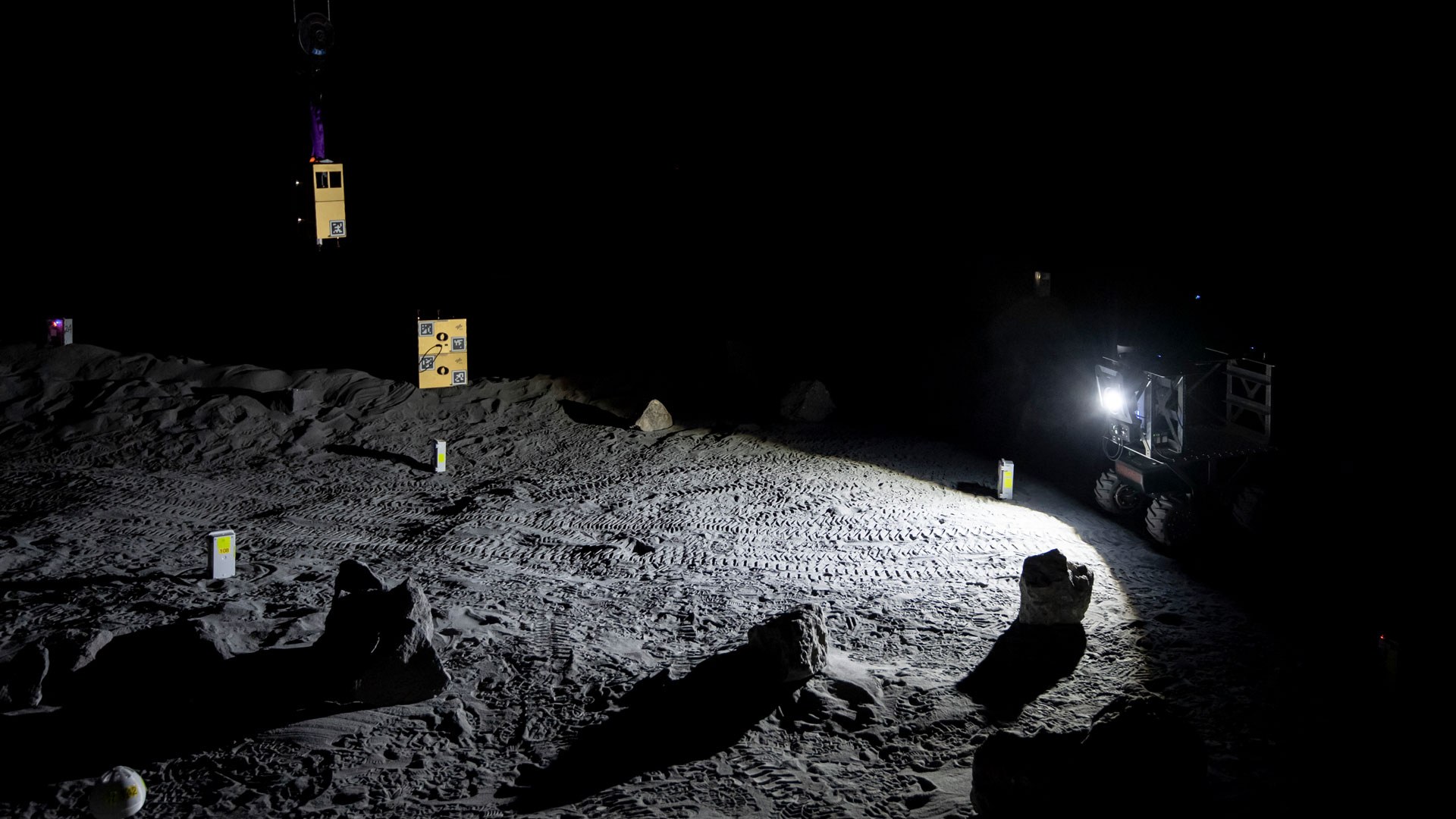 Das Bild zeigt einen Rover und Sensor-Einheiten in der dunklen LUNA-Halle. Die Lampe eines Rovers wirft einen Lichtkegel auf den grauen Regolith-Untergrund der LUNA-Halle.