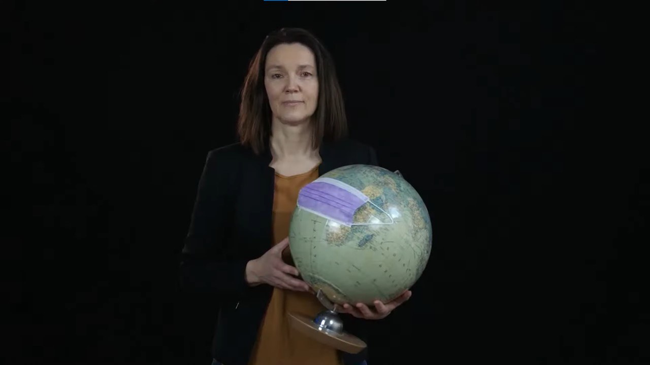 Zu sehen ist Dr. Claudia Nobis mit einem Globus in der Hand. Der Globus trägt eine OP-Maske. Anhand des Globus erklärt sie dem Zuschauenden etwas.