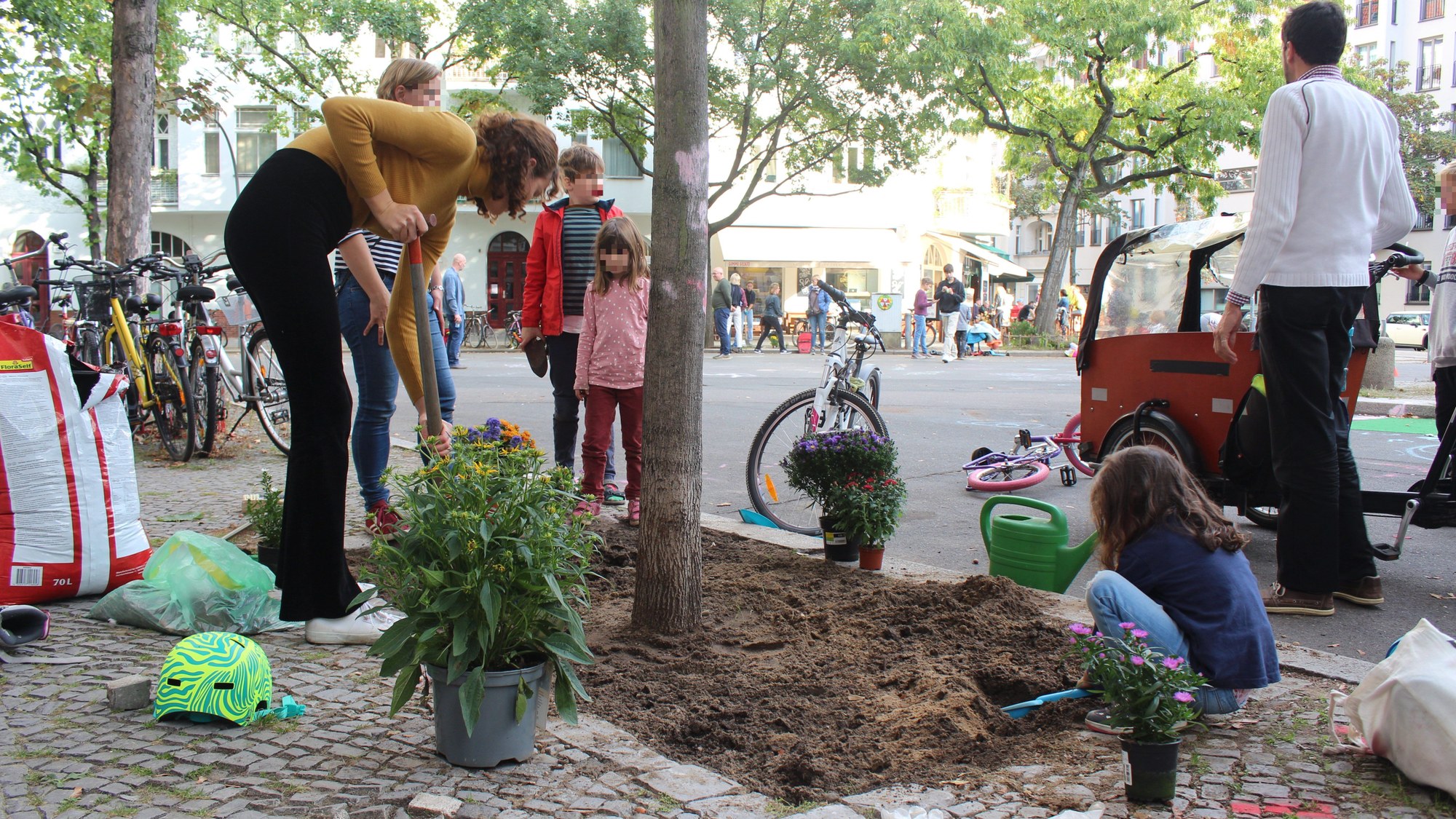 Zu sehen ist eine Gruppe von Kindern und Erwachsenen, welche gemeinsam eine Baumscheibe am Straßenrand eines verkehrsfreien Bereiches aufgraben, um ihn anschließend zu begrünen.