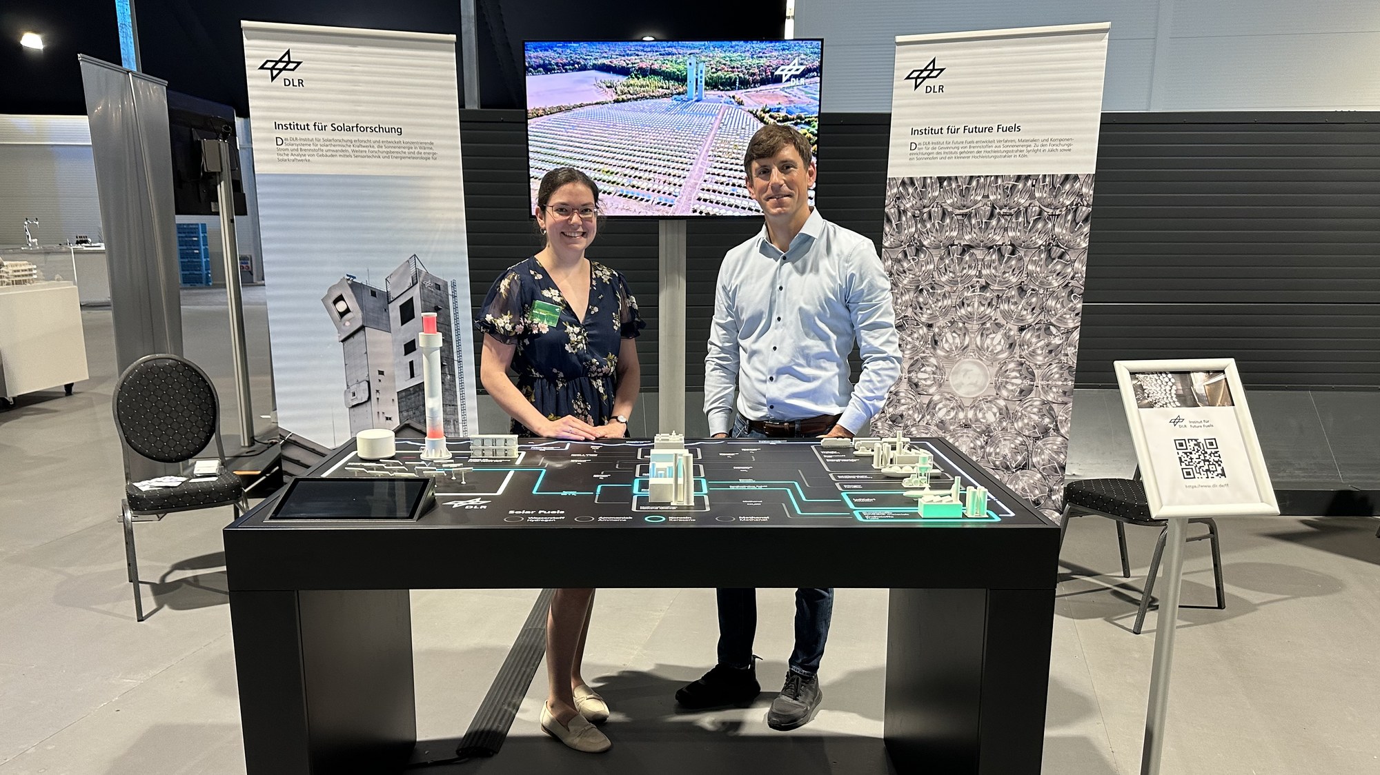 Two Researchers from the Institute of Future Fuels stand behind the Solar Fuels exhibit at DLR's stand.