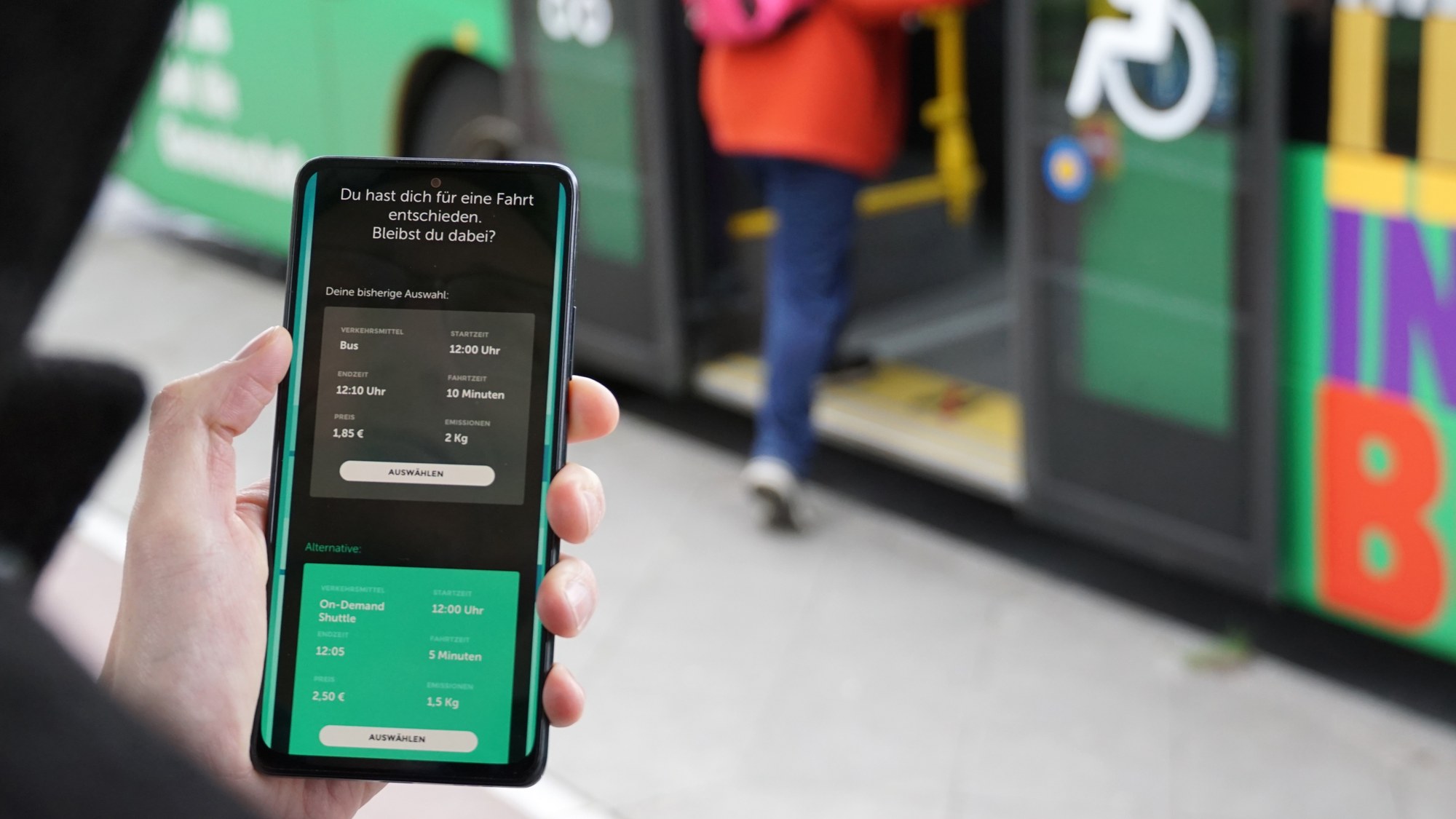 A Berlin BVG bus with green stickers and open doors with signs for wheelchairs and pushchairs in white can be seen blurred in the background. A person is boarding the bus. In the foreground, someone is holding a phone. The app is open on the phone. The screen is currently showing two options to choose from. Under the white lettering You have decided on a journey - if you stick with it, there are two boxes one below the other. One box in grey contains the bus offer with the properties travel time, price, emissions, etc. The second box in turquoise green describes the on-demand bus offer with the same properties. In both boxes there is an area for selecting the option.