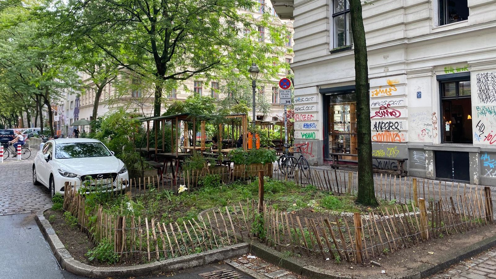 One of the redesigned parking bays on Böckhstraße. The border area was unsealed and greened. In the background, a car is parked, and the outdoor seating of a restaurant at the corner of the intersection can be seen.