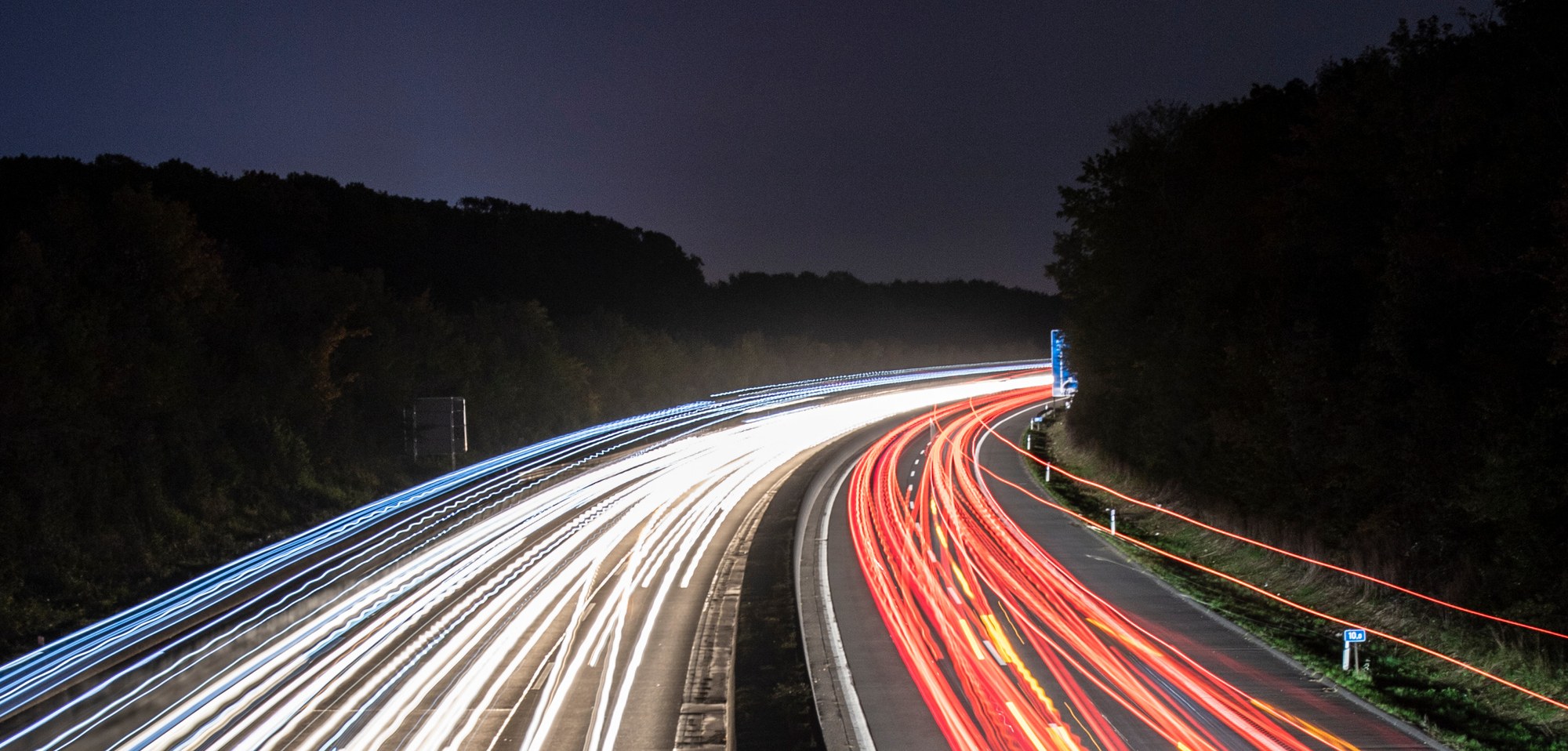 A motorway in the woods at night. Due to the long exposure time, the light from the headlights and tail lights of the vehicles has left bright white and red stripes on the lanes. These light strips appear to float above the motorway without any visible vehicles.